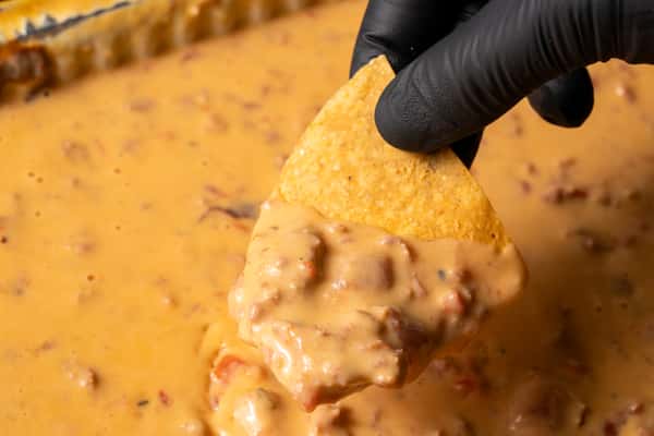 A hand dipping a tortilla chip into a pan of queso