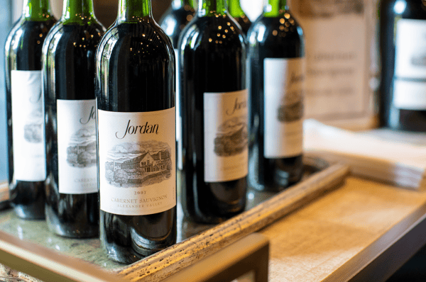 Close-up image of several bottles of Jordan Cabernet Sauvignon wine displayed upright on a table. The bottles feature cream-colored labels with an illustration of a winery estate and the text ‘Jordan Cabernet Sauvignon, Alexander Valley.’ The lighting reflects softly off the glass, highlighting the elegant presentation
