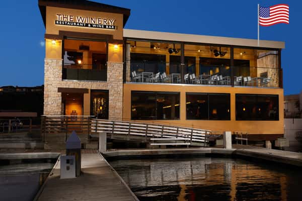 The Winery Restaurant & Wine Bar in Newport Beach at dusk, featuring a two-story waterfront building with a modern stone-and-stucco exterior, illuminated signage, expansive second-floor patio with glass railings, and a private dock overlooking the marina. An American flag is displayed on the upper right side of the building.
