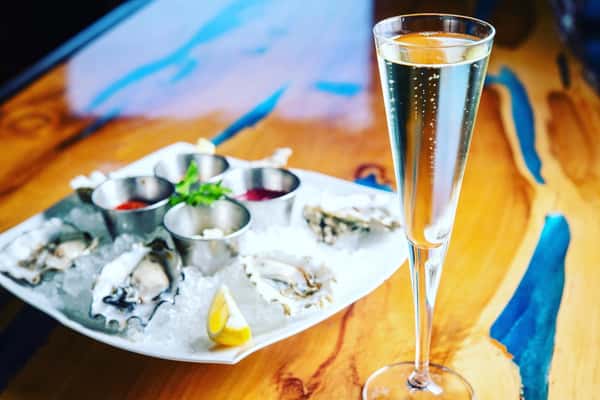 6 oysters on the half shell with an assortment on sauces on ice on a plate with a glass of champagne on a brown and blue table top