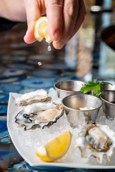 fresh shucked oysters on ice with lemon and sauces with a hand squeezing lemon over the oysters. plate on blue agate.