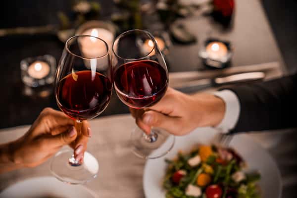 Close-up of two guests clinking glasses of red wine at a candlelit restaurant table with plated food in the background.