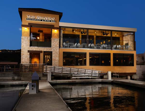 The Winery Restaurant & Wine Bar Newport Beach is shown at dusk on the waterfront. The two-story restaurant sits directly above the marina and features warm architectural lighting, large glass windows, stone accents, and a spacious second-story patio with harbor views. A dock and walkway lead guests to the entrance, while the restaurant's lights reflect across the water, creating an inviting coastal dining atmosphere.