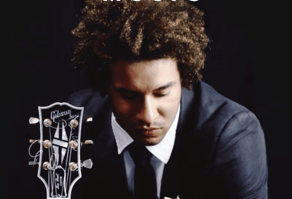 A well-dressed male musician with curly brown hair, wearing a black suit, white shirt, and black tie, sits with his head slightly bowed and eyes closed in a reflective pose. In front of him is the headstock of a black Gibson Les Paul guitar, with silver tuning pegs and logo clearly visible. The background is dark, emphasizing the artist and guitar. The mood is introspective and artistic.