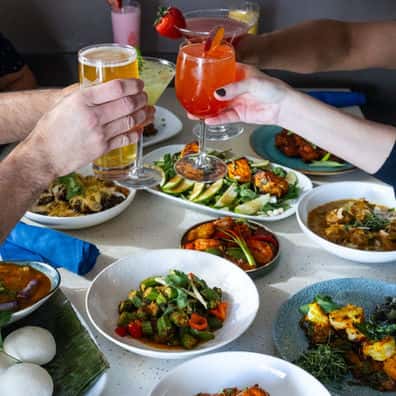 Two hands cheering cocktails over a spread of Indian food from Rasoi
