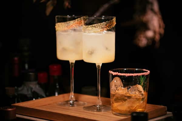 Two tall glasses of citrus cocktails with gold-rimmed edges beside a short glass of whiskey on ice with a pink sugar rim.
