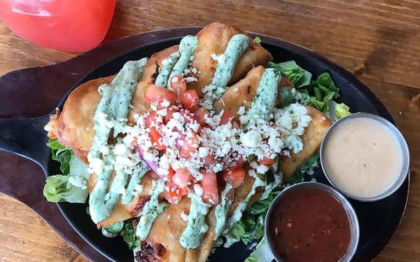 Crispy steak taquitos on a skillet over lettuce with spicy green sauce, salsa and sour cream with a strawberry margarita