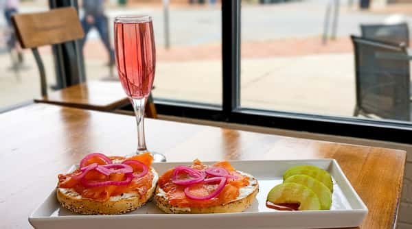 Lox bagels with cream cheese and red onion, sliced green apples with syrup, and a pink drink in a flute glass.