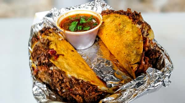 Two birria tacos with melted cheese and a side of red consommé with cilantro, served in an aluminum foil tray.