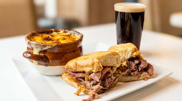 French onion soup with melted cheese, steak sandwiches, and a glass of dark beer on a white plate.