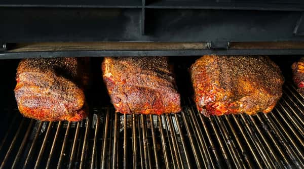 Three large cuts of BBQ meat, seasoned with black pepper and spices, cooking on a dark metal grill inside a smoker.