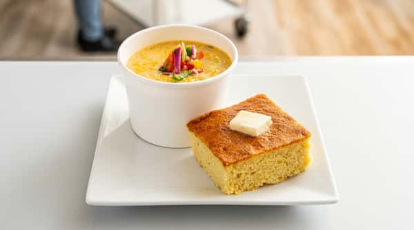 Corn chowder with colorful diced vegetables in a white cup and a square of cornbread with butter.