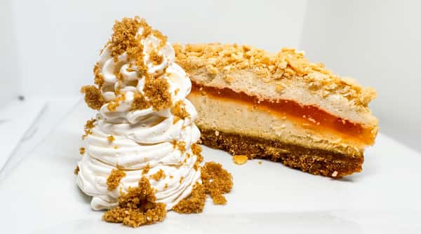 Cheesecake slice with an orange jam layer and crumbled topping, next to a dollop of whipped cream with brown sugar sprinkles.