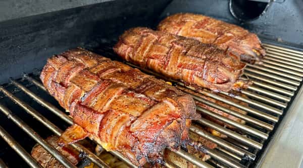 Three bacon-wrapped pork loins with seasoning cooking on a grill grate.