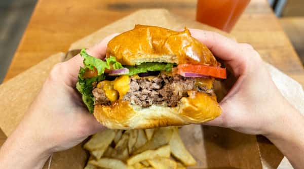 Hands holding a partially eaten cheeseburger with lettuce, tomato, and onion over a container of fries.