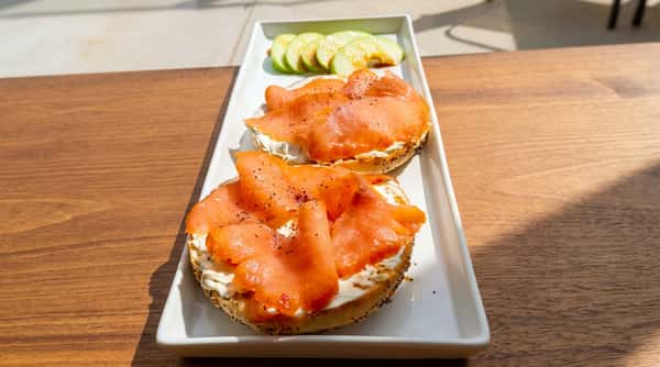 Two everything bagels with cream cheese and smoked salmon on a white rectangular plate, with sliced green apple and sauce.