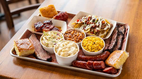 A BBQ platter with sliced brisket, sausage, burnt ends, nachos, coleslaw, mac and cheese, corn, baked beans, and cornbread squares.