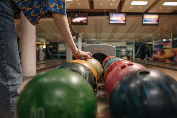 Person reaching for a green bowling ball from a return rack at a bowling alley, with colorful balls lined up.
