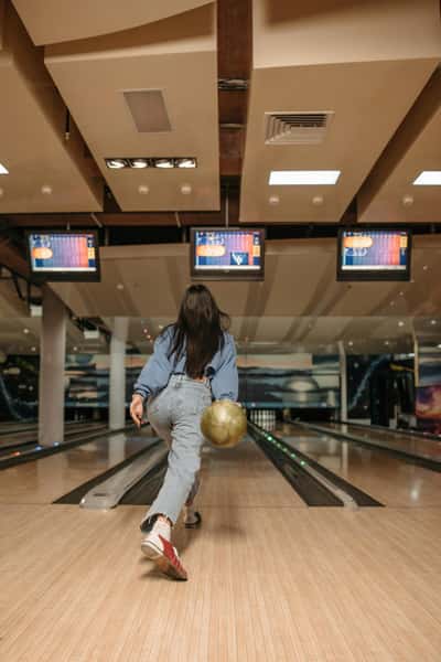 Woman in jeans and red sneakers bowling, holding a golden ball while approaching the lane under overhead scoring screens.