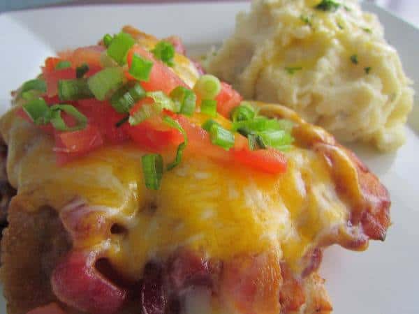 Close up image of a burger with bacon, melted cheddar cheese and diced tomato with scallions. Served with a side of mashed potatoes,