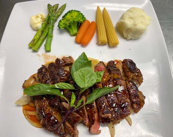 Grilled sliced steak served with mashed potatoes, baby corn, carrots, broccoli, asparagus, and a dollop of sauce on a white plate