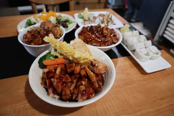 A bowl filled with grilled chicken, tempura vegetables, and rice, with other Japanese dishes blurred in the background