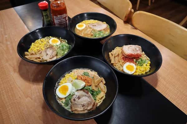 "Four bowls of ramen with sliced pork, soft-boiled eggs, corn, green onions, and bamboo shoots, served on a wooden table with condiments