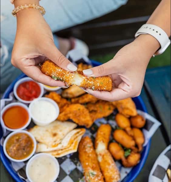 Close-up of hands pulling apart a crispy mozzarella stick with gooey melted cheese, above a shareable appetizer platter featuring fried chicken tenders, mini corn dogs, quesadilla wedges, and assorted dipping sauces—perfect for bar snacks or game day bites.