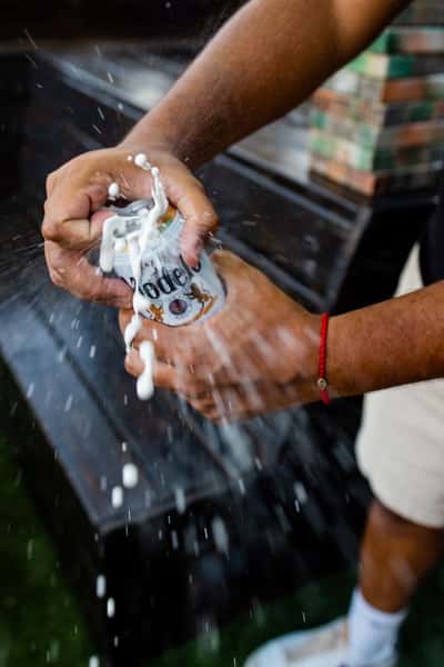 Close-up of a person opening a Modelo beer can as it explosively foams over, capturing a dynamic and energetic moment typical of a lively bar or tailgate atmosphere.