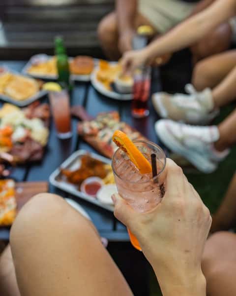 Person holding a refreshing cocktail garnished with an orange slice during an outdoor gathering, with a table full of bar food favorites like flatbread pizza, chicken wings, fries, sliders, and charcuterie in the background—perfect for a social brunch or weekend patio hangout.
