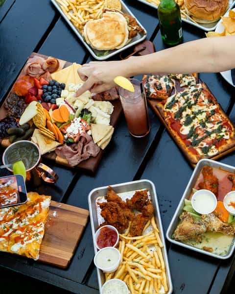 Overhead view of a vibrant outdoor dining table filled with a variety of American bar food, including a loaded charcuterie board with cured meats, cheeses, fresh fruits, crackers, and pickles, surrounded by crispy chicken wings, French fries, buffalo chicken pizza, fried chicken sandwiches, and a rectangular flatbread pizza with melted mozzarella and basil. A hand reaches for cheese on the charcuterie board, with cocktails and a beer bottle completing the lively, casual dining scene.