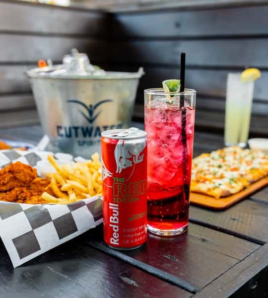 Vibrant Red Bull vodka cocktail made with Red Bull Red Edition and garnished with a lime wedge, placed beside crispy chicken tenders, French fries, and a flatbread pizza, with a bucket of canned cocktails in the background—ideal for energetic patio dining or weekend happy hour.