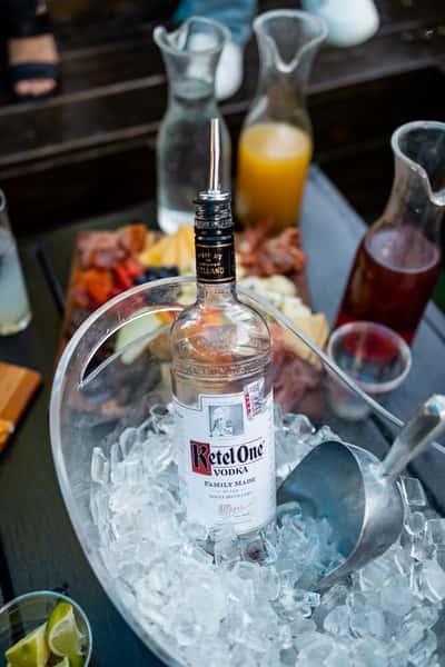 Ketel One vodka bottle chilling in a clear ice bucket with a metal scoop, surrounded by cocktail mixers in glass carafes and a charcuterie board with assorted meats, cheeses, and fruit—ideal setup for a premium outdoor brunch or DIY cocktail experience.