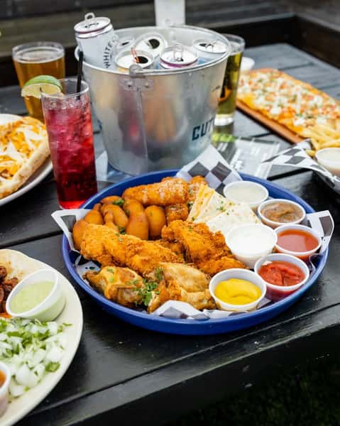 Large platter of assorted bar bites including crispy chicken tenders, fried mozzarella sticks, soft pretzel bites, and quesadilla slices, surrounded by a variety of dipping sauces. The table also features a vibrant red cocktail, beer, and a metal bucket filled with canned seltzers on ice, perfect for a casual outdoor dining experience.