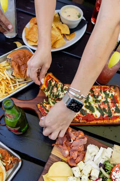 Person grabbing a slice of cheesy flatbread pizza topped with pesto and tomato sauce, surrounded by loaded plates including a charcuterie board with assorted meats and cheeses, French fries, a burger, and chips with queso—perfect for a lively group dining experience.