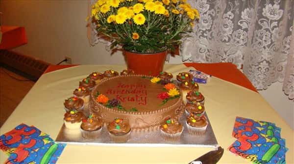 Chocolate birthday cake decorated with chocolate frosting and colorful flower rosettes surrounded with chocolate cupcakes on a table wih colorful napkins and a small flowerpot with yellow flowers