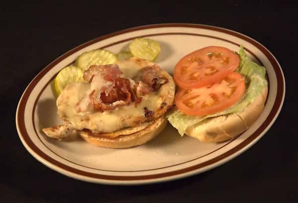 Chicken On The Ranch Sandwich, open faced and topped with bacon, Swiss cheese, tomato and lettuce. Served with slices of dill pickle.