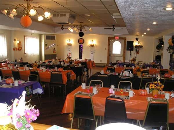 Dining hall with tables set for Halloween in orange and purple linen, tables decorated with pumpkins and and fall flowers and balloons and flower decorations on the wall