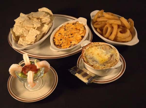 Variety of plates with appetizers. Chicken wing dip with tortilla chips in a platter next to a plate of fried onion rings, a tall glass of shrimp cocktail and soup with melted cheese in a bowl