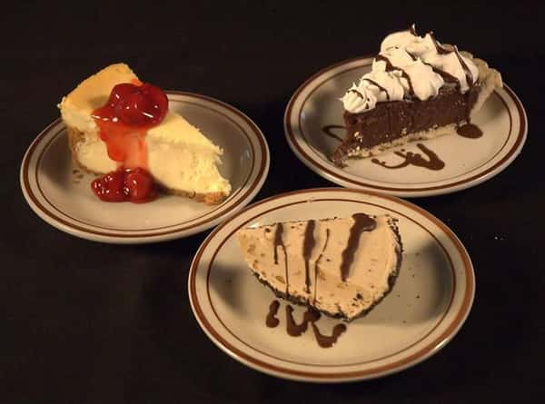 Variety of small plates with pies and cakes. A piece of cheesecake topped with cherries, a plate of peanut butter pie and a plate of chocolate cream pie