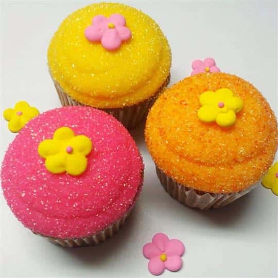 Three cupcakes with different frostings (yellow, pink, and onange) topped with sugar flowers