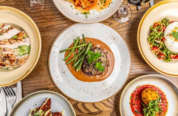 Herb-crusted steak with green beans in brown sauce, surrounded by grilled chicken, pasta, burrata salad, and arancini.