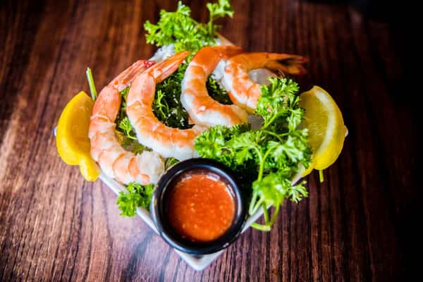 A beautifully plated shrimp cocktail, arranged over fresh greens with lemon wedges and a side of tangy dipping sauce.