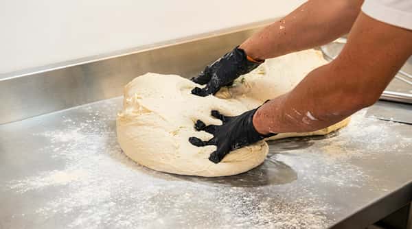 Hands in black gloves kneading a large mound of white dough on a gray, floured counter.