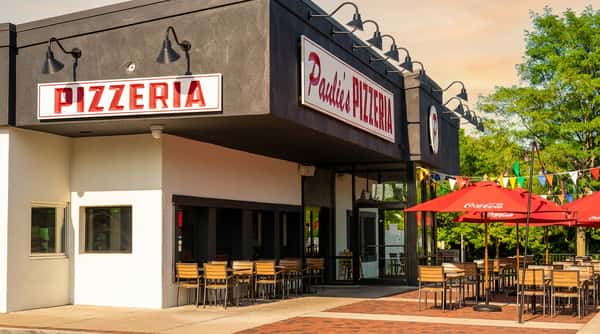 Exterior of Paulie's Pizzeria with white signs featuring red lettering, black awning, and red umbrellas.