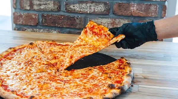 Hand in a black glove lifting a slice of cheese pizza from a large round pizza.