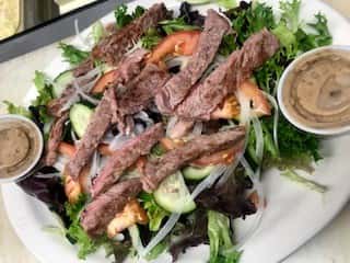 Steak salad with mixed greens, tomatoes, cucumbers, onions, and grilled steak strips, served with two cups of dressing.