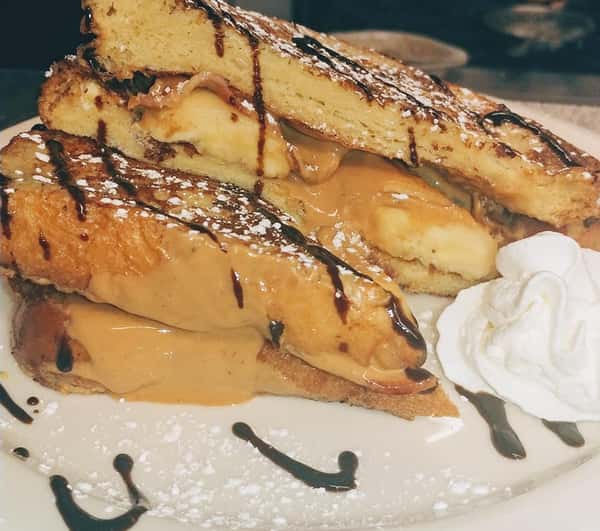 Thick slices of French toast stuffed with creamy peanut butter and banana slices, drizzled with chocolate syrup and dusted with powdered sugar, served with a dollop of whipped cream.