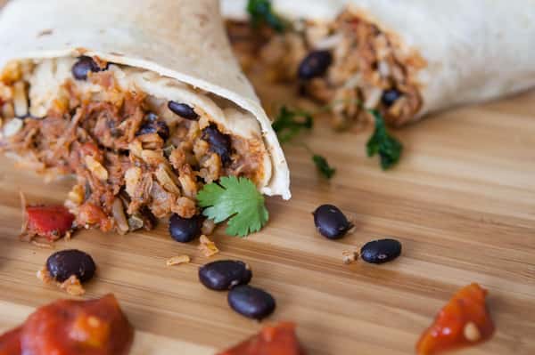 Burrito filled with shredded meat, rice, and black beans on a wooden board with scattered cilantro, salsa, and beans.