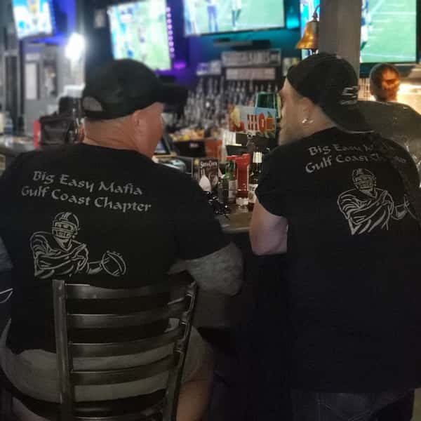 Two patrons seated at a sports bar counter wearing matching football-themed shirts, drinks and condiments on the bar, with TVs showing live games in a lively American pub atmosphere.